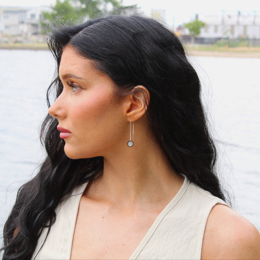 Woman with long dark hair and earrings standing by a body of water with buildings in the background