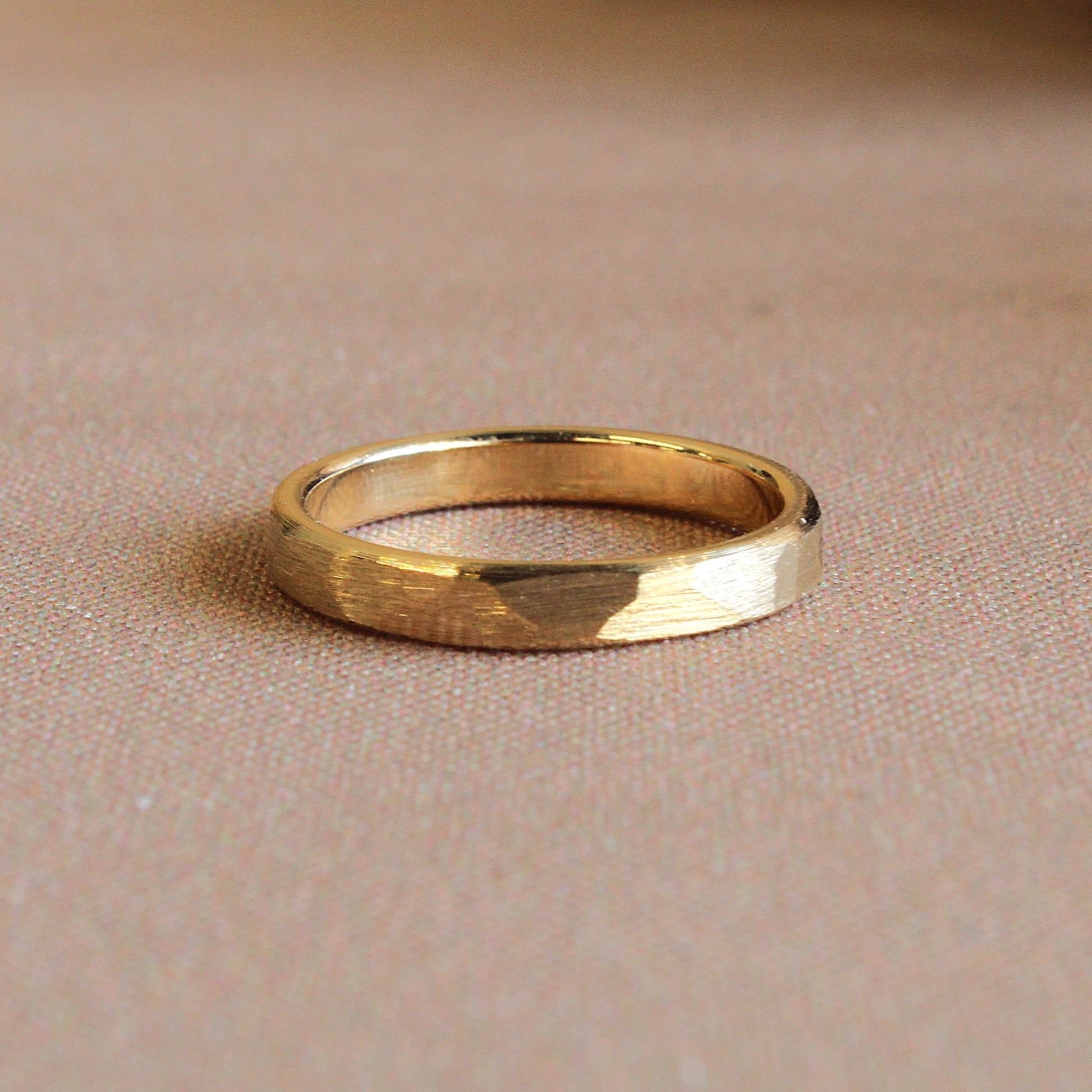 A gold band ring with a matte finish and slight texture, displayed on a beige background.