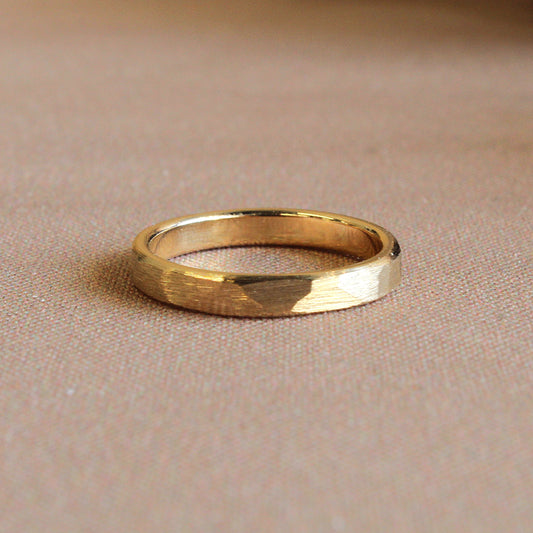 A gold band ring with a matte finish and slight texture, displayed on a beige background.