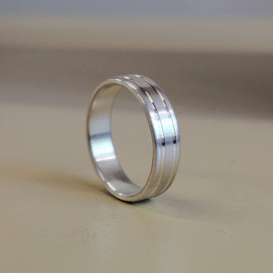 A silver metal band ring with a polished finish and subtle linear grooves, standing upright on a neutral beige surface.