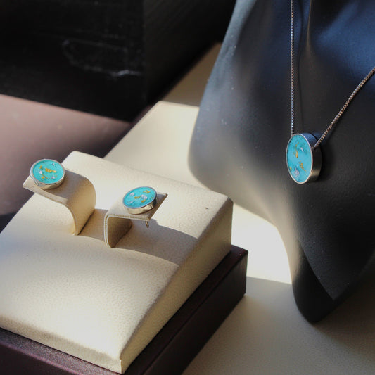 A jewelry display showing a pair of oval stud earrings in a cream jewelry box next to a black mannequin bust wearing a matching necklace. The oval studs feature a bright turquoise blue center with small gold details, set in silver-toned metal.