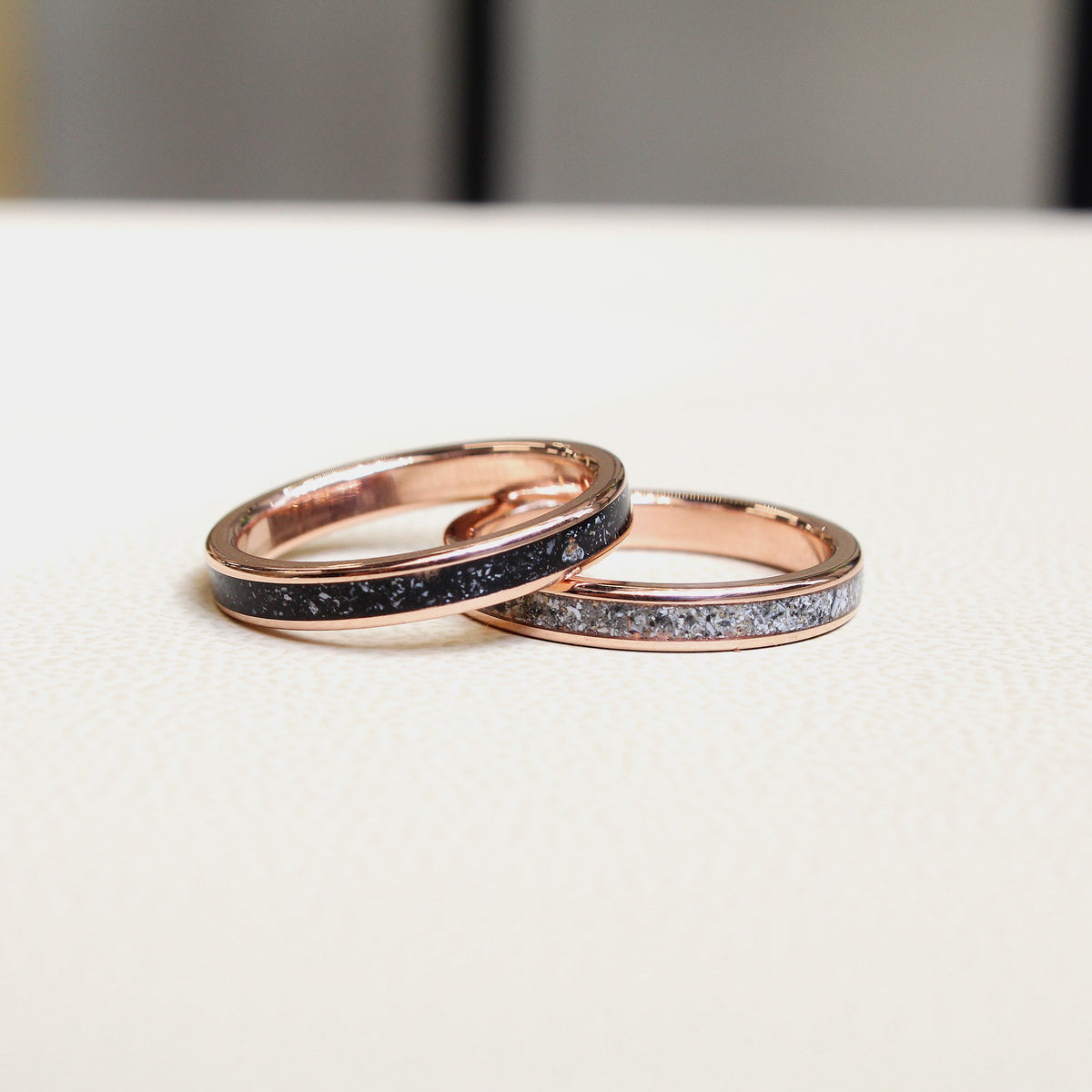 Two rose gold rings with black and silver ashes, resting on a light beige textured surface.