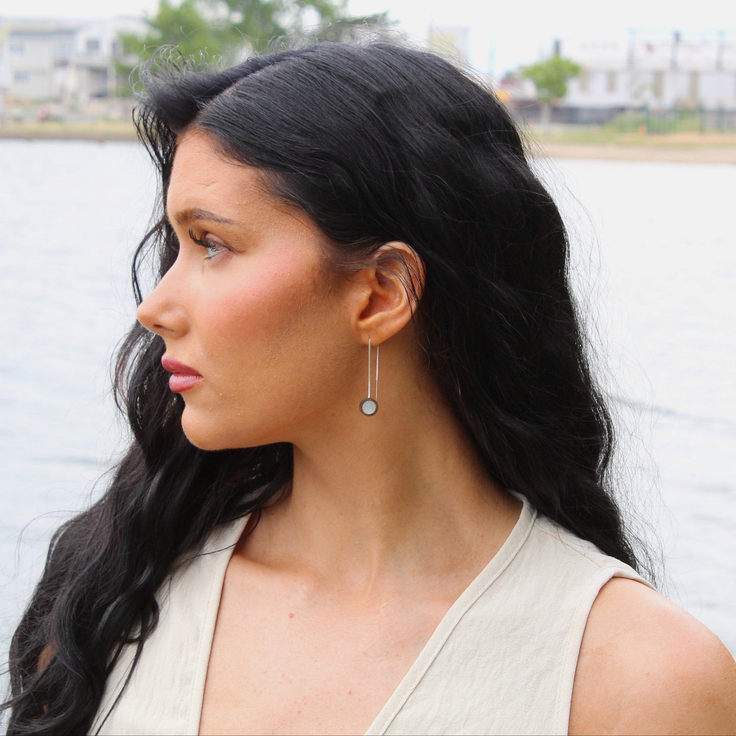Woman with long dark hair and earrings standing by a body of water with buildings in the background