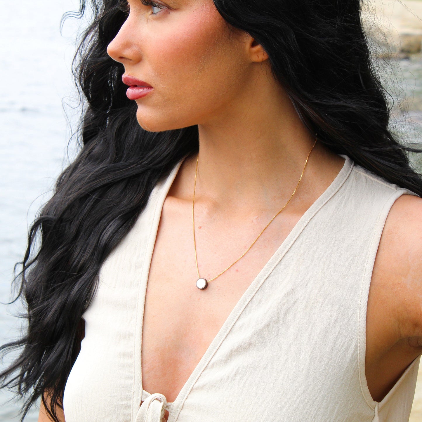Woman with long dark hair wearing a white sleeveless top and gold necklace by a water body.
