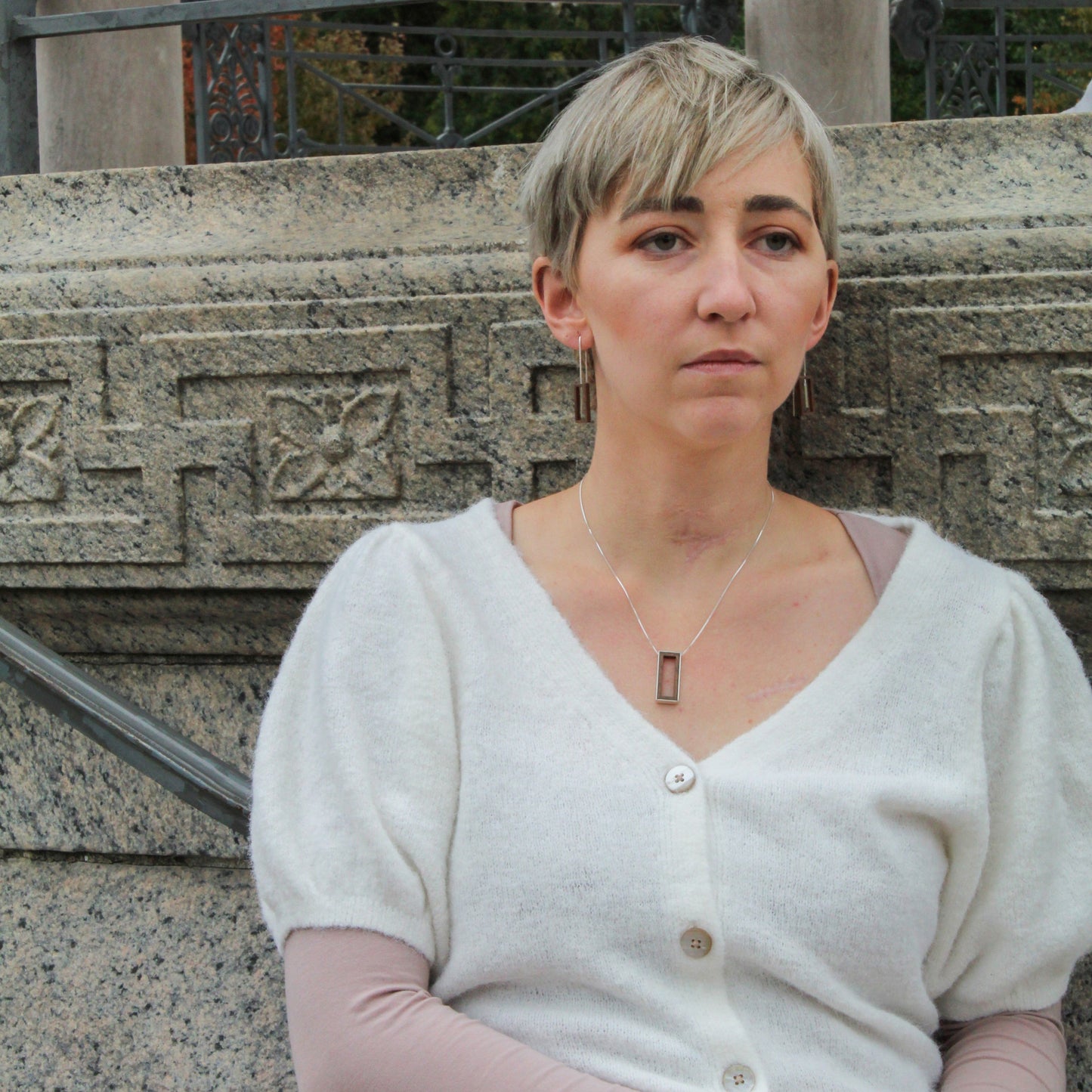 Woman standing outdoors with a stone wall and greenery in the background wearing wood and silver jewelry 