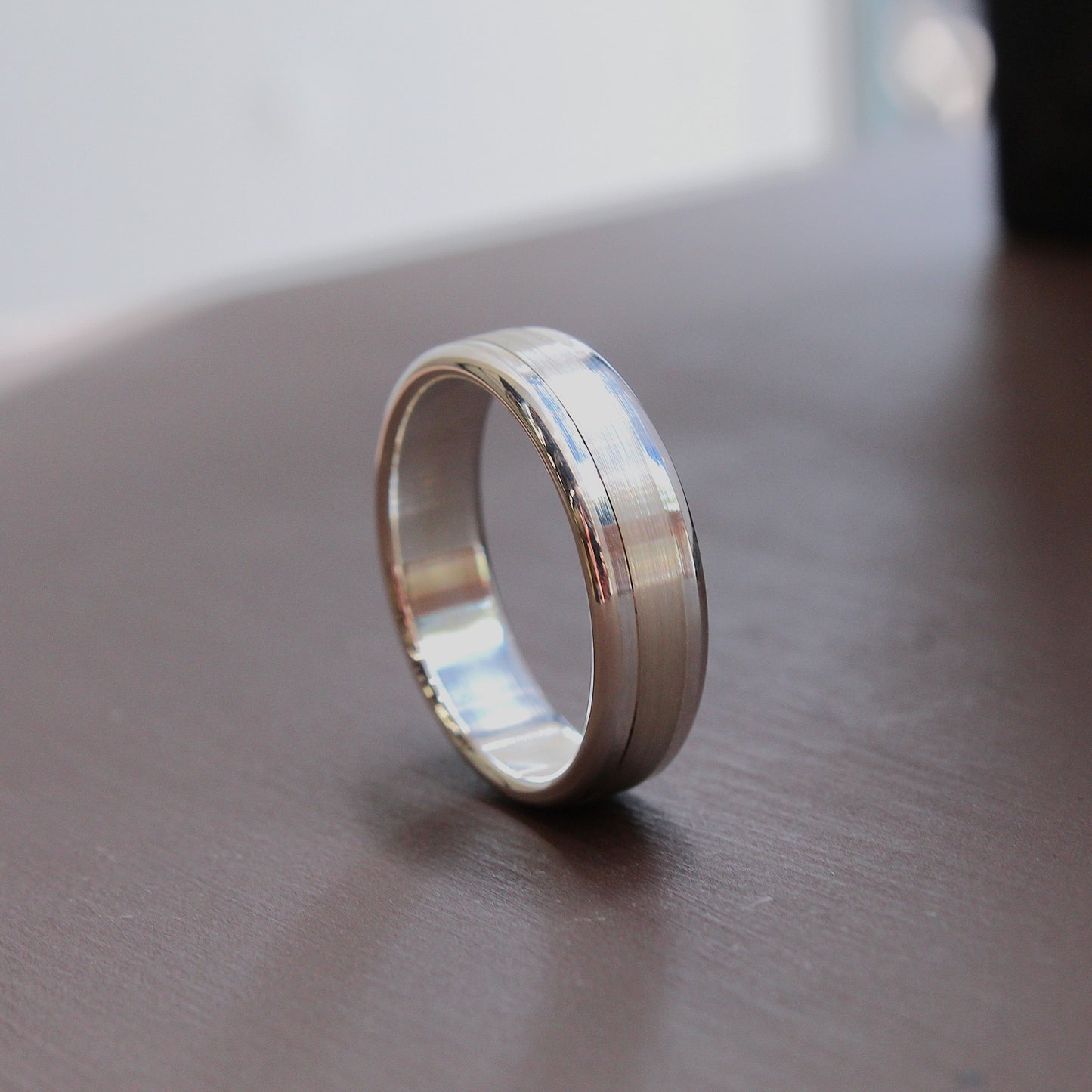 A silver-toned metal band ring standing upright on a dark brown wooden surface, with a softly blurred light gray background.