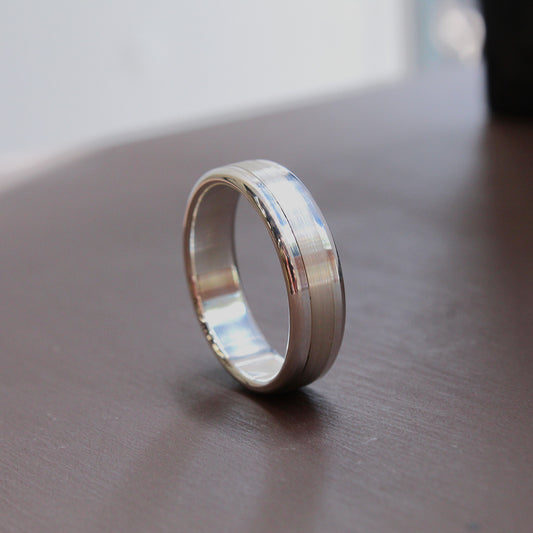 A silver-toned metal band ring standing upright on a dark brown wooden surface, with a softly blurred light gray background.