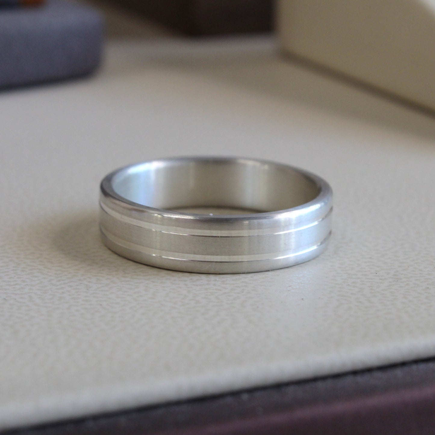 A silver-toned metal band ring with a textured, brushed center channel and polished beveled edges, resting on a light beige textured surface.