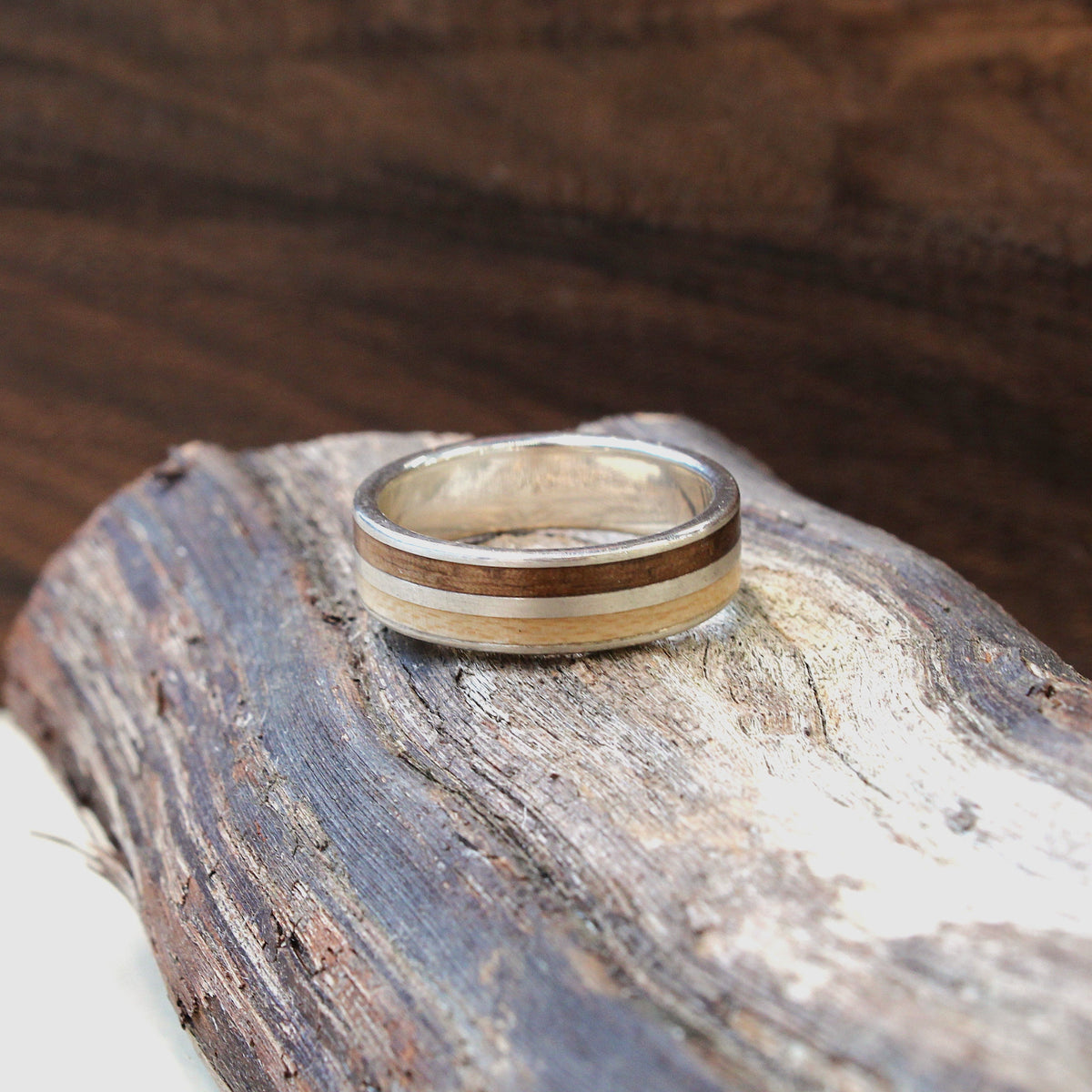A silver-toned ring with two horizontal wood inlays, one dark brown and one light beige, resting on a piece of weathered driftwood with a dark wood background.