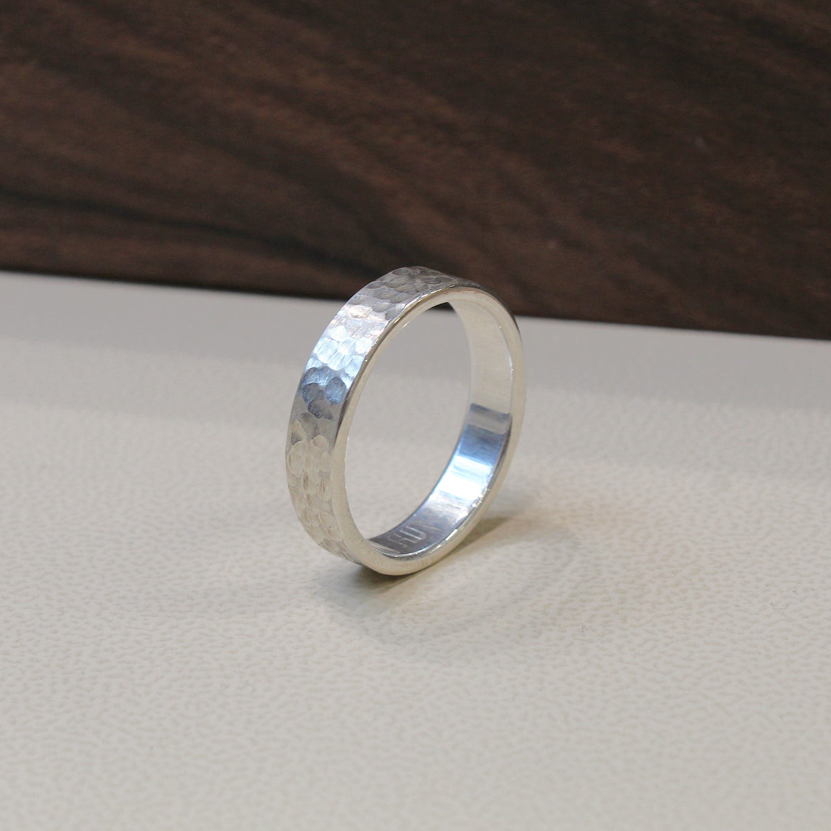 A hammered silver band ring standing upright on a light beige textured surface with a dark wood background.