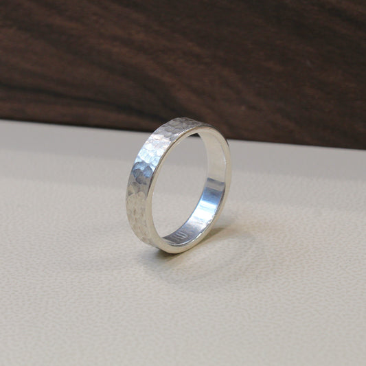 A hammered silver band ring standing upright on a light beige textured surface with a dark wood background.