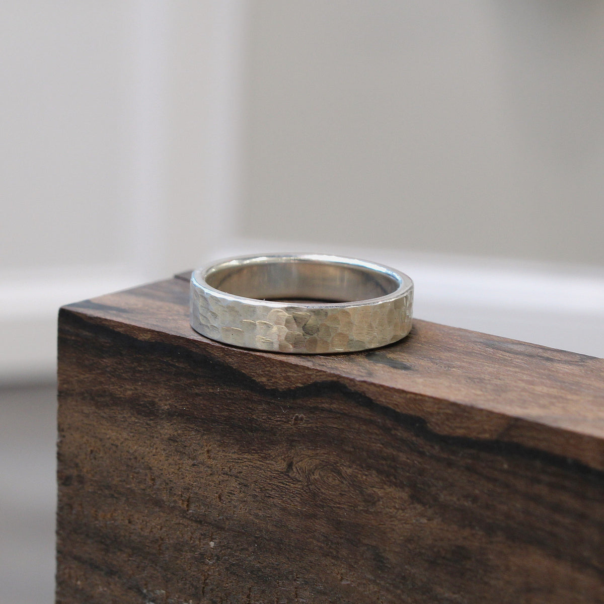 A hammered silver band ring resting on the corner of a dark wood block, with a softly blurred indoor background.