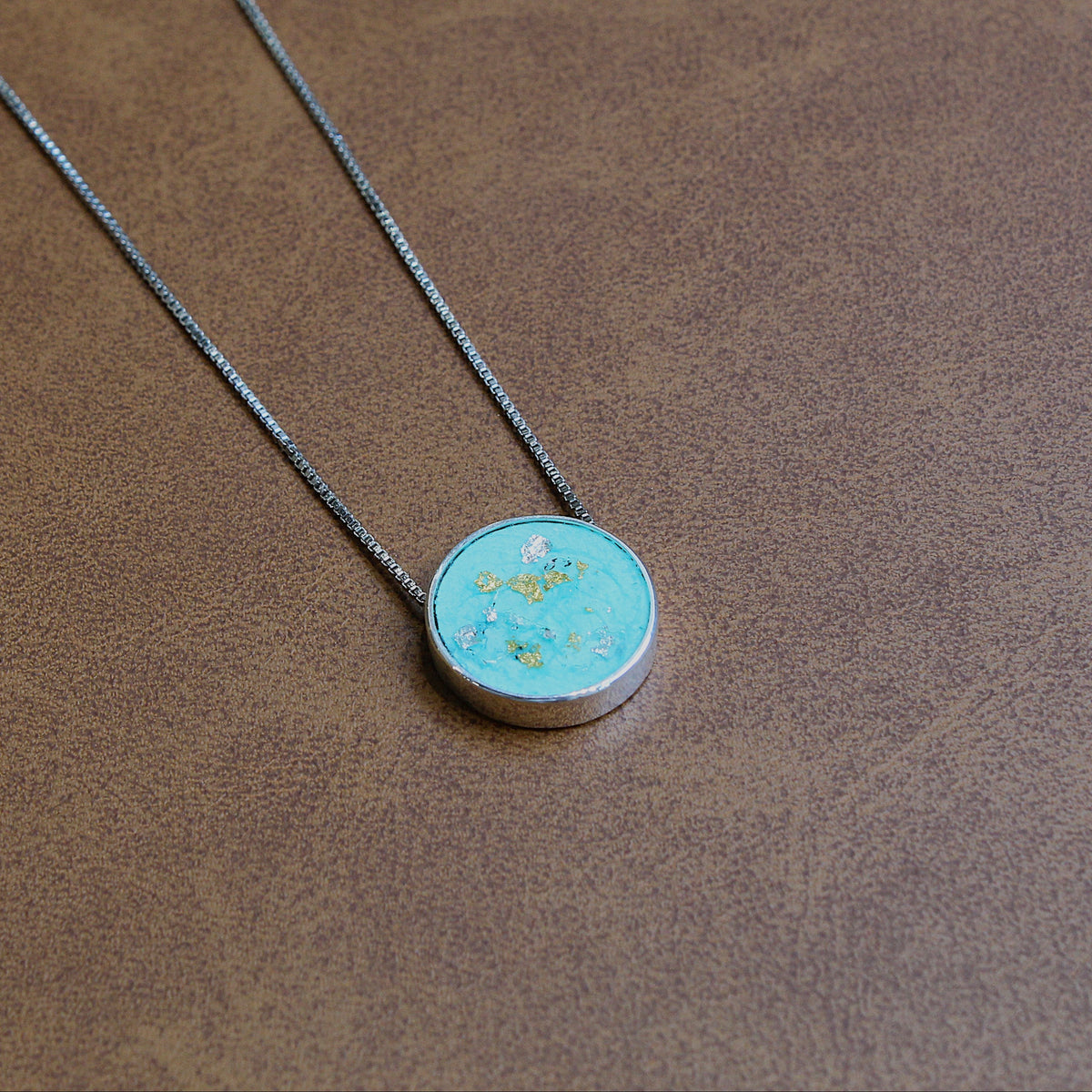 A silver chain necklace with a round pendant featuring a light blue-green turquoise-like center with small gold flecks, displayed on a textured brown surface.