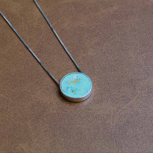 A silver chain necklace with a round pendant featuring a light blue-green turquoise-like center with small gold flecks, displayed on a textured brown surface.