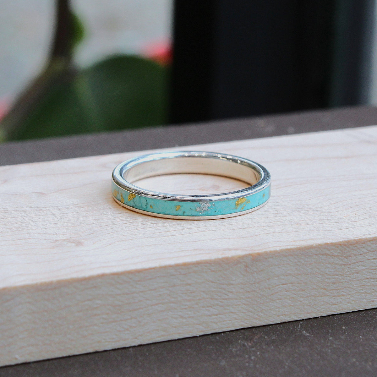A thin silver band ring with a turquoise inlay rests on a light-colored wooden block, with a blurred background showing green leaves and a dark surface.