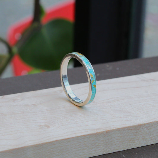 A silver band ring with a small turquoise inlay, standing upright on a light-colored wooden block with a blurred background.